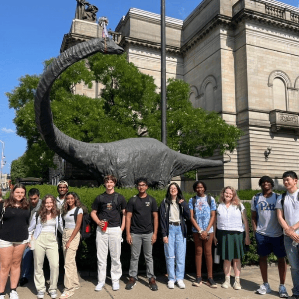 Group of students stand in front of dinosaur statue in Oakland.
